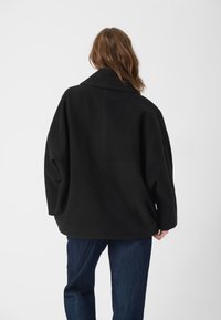 Musta ylisuuri takki, jossa on jämäkkä kaulus, valmistettu sileästä kankaasta. Ominaisuuksiin kuuluvat roikkuneet olkapäät ja puhdas, minimalistinen muotoilu.