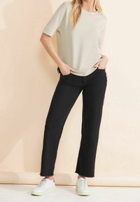 Světlé béžové tričko s krátkými rukávy v kombinaci s černými džíny s vysokým pasem a neupraveným lemem. Nošeno s bílými teniskami na jemném pozadí.