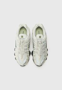 Nike Shox sneakers i ljusgrått mesh med texturerade accenter, har framträdande sula med stötdämpning och ett distinkt snörningssystem.