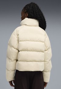 Krämfärgad pufferjacka med en distinkt kordflorsstruktur, hög krage och en kortad silhuett, med horisontellt quiltade sektioner.