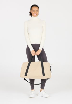 Sac de voyage beige avec des sangles noires, fabriqué en tissu lisse. De grande forme rectangulaire, il dispose d'une bandoulière amovible et d'une fermeture éclair.