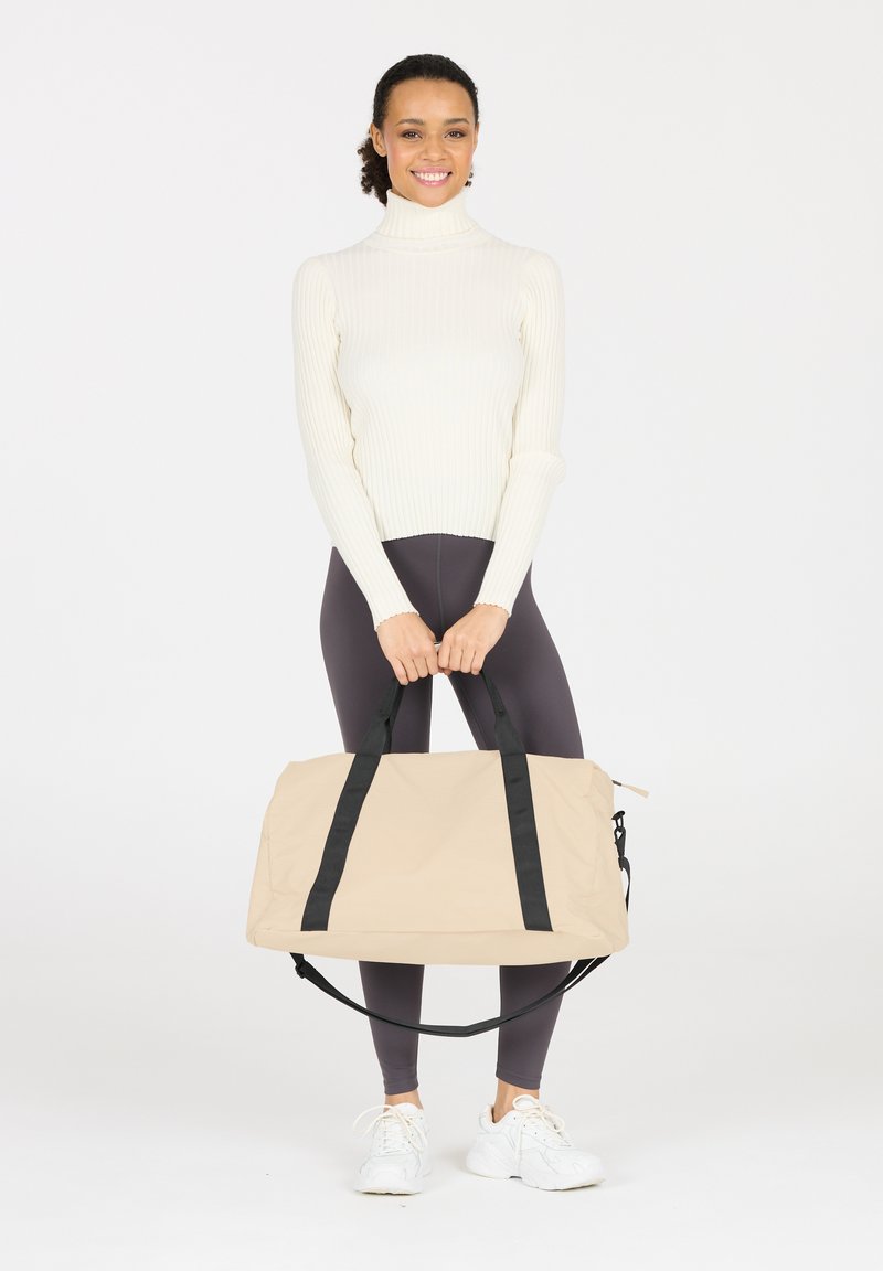 Bolsa de viaje beige con correas negras, hecha de tela suave. Forma rectangular grande, con una correa para el hombro desmontable y cierre de cremallera.