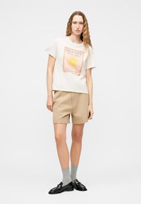 Γυναίκα με μακριά κυματιστά μαλλιά, που φοράει λευκό T-shirt με γραφικά, μπεζ σορτς, γκρι κάλτσες και μαύρα μοκασίνια, στέκεται μπροστά σε απλή φόντο.