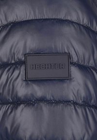 Marineblaues Steppjacke mit strukturiertem Design, versehen mit einem rechteckigen schwarzen Etikett, das das Wort "HECHTER" geprägt für die Markenidentität trägt.
