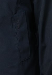 Casaco azul marinho com um tecido texturizado. Apresenta um bolso lateral com o logótipo "Barbour" em relevo. Design limpo e clássico com acentos mínimos.