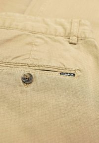 Khakifarbene Stoffhosen mit einer Gesäßtasche mit Knopfverschluss und einem kleinen Markenlabel, präsentiert vor einem strukturierten Hintergrund.