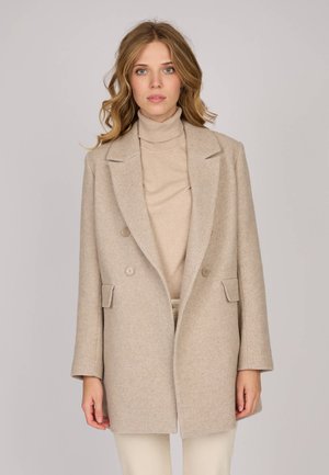 Femme avec des cheveux blonds ondulés portant un manteau en laine beige à double boutonnage sur un pull à col roulé assorti et un pantalon de couleur claire.