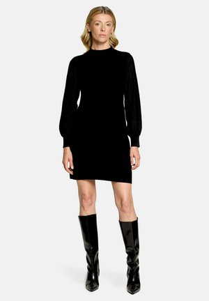 Femme portant une mini-robe noire à manches longues bouffantes et des bottes noires brillantes montant jusqu'aux genoux, debout devant un fond clair uni.