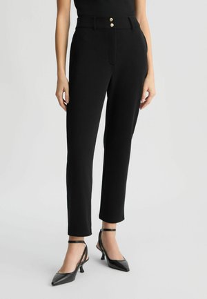 Pantaloni neri aderenti con una texture liscia, dotati di vita alta, chiusura con due bottoni dorati e gambe affusolate. Indossati con tacchi neri a punta.