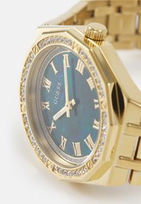 Gouden octagonale horloge met een blauw iriserend uurwerk, Romeinse cijfermarkeringen, een bezel met kristallen accent en een metalen schakelarmband.