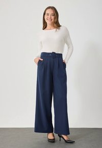 Marinblå vida byxor med bälte i midjan och veck. Kombinerade med en figurnära vit långärmad topp och svarta högklackade skor.