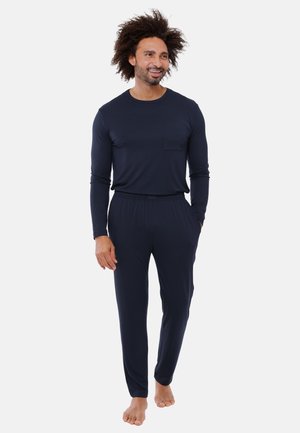 Uomo con capelli ricci che indossa una camicia blu navy a maniche lunghe e pantaloni coordinati, in piedi a piedi nudi con una mano in tasca.