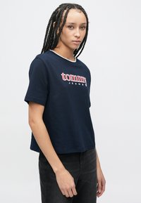 Tengerészkék pamut póló fehér gallérral, piros és fehér "tommy" logóval. Rövid ujjú, laza szabású. Fekete farmernadrág van rajta.