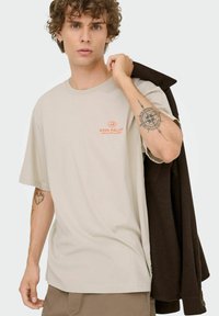 Camiseta de algodón beige con un corte relajado, que presenta el texto naranja "ROSA PÁLIDO" en el lado izquierdo del pecho. Modelo llevando una chaqueta marrón oscuro sobre el hombro.