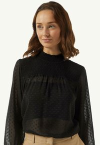 Schwarze transparente Bluse mit strukturierten Punkten, gerafftem hohen Kragen und langen Ärmeln. Der Stoff ist leicht und fließend und hat einen sanften Fall.