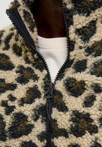 Fleecejakke med et leopardprint mønster i beige og mørkegrøn, med lynlås fortil og struktureret stof. Sort lynlås bånd og træk.