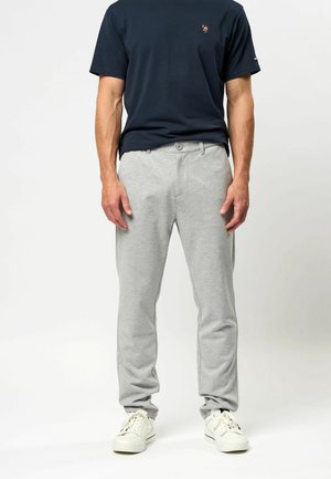 Mężczyzna ubrany w granatowy t-shirt, jasnoszare spodnie casualowe i białe trampki, stojący na tle gładkiej białej ściany.