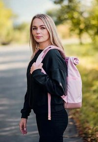 Růžový látkový batoh s rolovacím uzávěrem, nastavitelnými popruhy a texturovaným povrchem. Nosený na černé mikině, umístěný v přírodním prostředí.