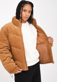 Ruskea corduroy-toppatakki, jossa on timantin muotoinen tikkauskuvio, korkea kaulus ja vetoketjusuljenta. Sisävuori on vaaleanbeige ja siinä on tasku.
