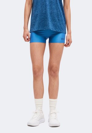Személy kék sport rövidnadrágot visel Under Armour logóval, kék pólót, fehér zoknit és fehér sportcipőt, egyszínű háttér előtt áll.