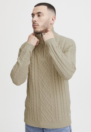 Beżowy sweter dzianinowy z kołnierzem na pół-zamek, z wzorem w warkocze na klatce piersiowej i rękawach oraz ściągaczami przy mankietach i u dołu.