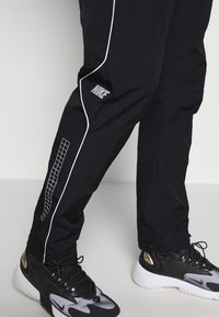 Svarta Nike träningsbyxor med vit piping och ett rutmönster nära ankeln, tillsammans med svarta och vita Nike sneakers med tjocka sulor.