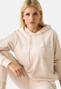 Béžová mikina z měkké tkaniny, s kapucí na šňůrku, detailním logem z kamínků a volným střihem. Doplněná odpovídajícími kalhotami.