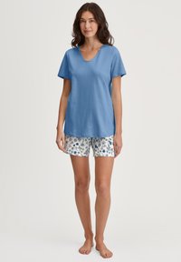 Blaues Baumwoll-V-Neck-T-Shirt, kombiniert mit weißen Shorts, die ein florales Muster in Blau und Grün aufweisen. Kurze Ärmel und lockere Passform.
