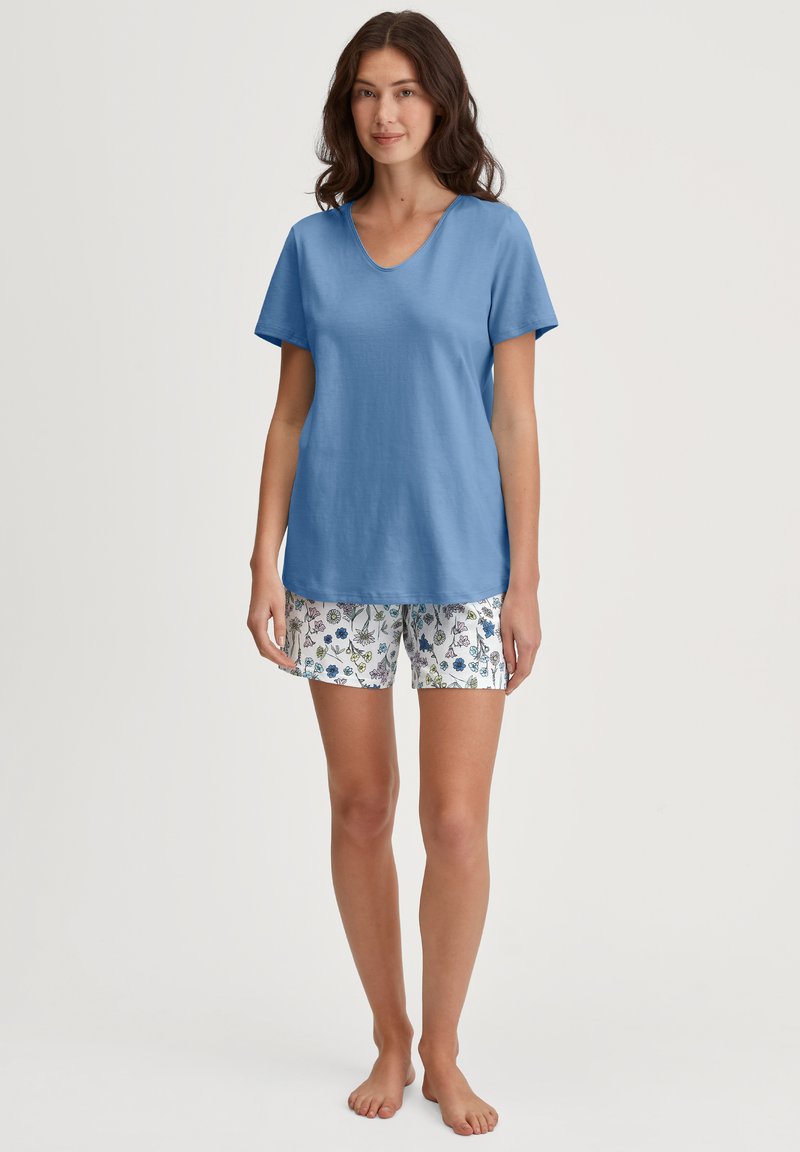 Blaues Baumwoll-V-Neck-T-Shirt, kombiniert mit weißen Shorts, die ein florales Muster in Blau und Grün aufweisen. Kurze Ärmel und lockere Passform.