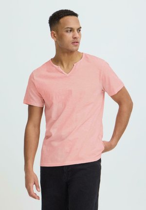 Jeune homme portant un t-shirt à manches courtes col en V rose clair et un pantalon noir, regardant vers sa gauche avec une main derrière le dos.