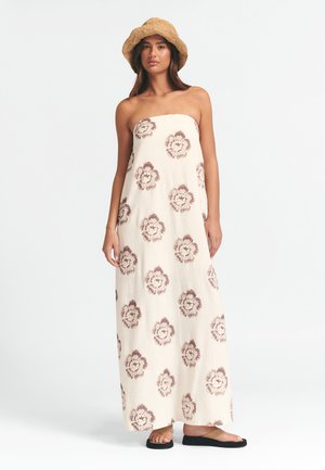 Stropeløs maxi kjole i lys beige stof med et blomsterprint i mørkerød. Båret med flade sorte sandaler og en vævet stråhat.
