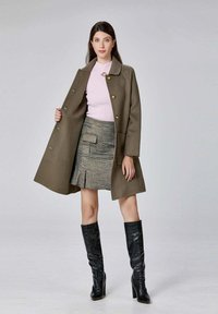 Manteau en laine vert olive avec des boutons dorés, associé à un pull rose clair et une mini-jupe en tartan. Bottes en cuir noires jusqu'aux genoux.