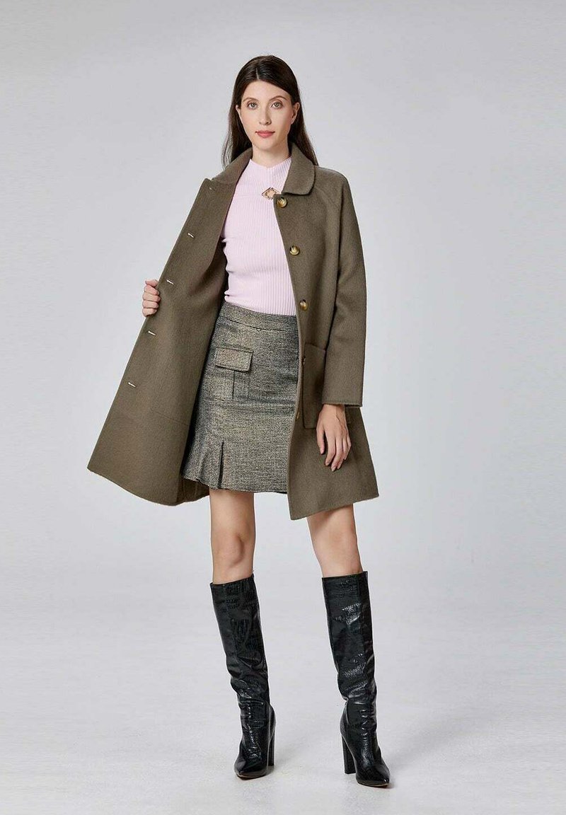 Manteau en laine vert olive avec des boutons dorés, associé à un pull rose clair et une mini-jupe en tartan. Bottes en cuir noires jusqu'aux genoux.