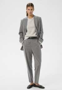 Giacca di suit grigia a righe sottili su un maglione chiaro, abbinata a pantaloni su misura. Cintura e scarpe nere completano il look. Tessuto liscio e strutturato.