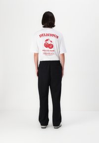 White t-shirt with red graphic of cherries and text on back, paired with black trousers. Simple design, short sleeves, casual fit.