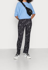 Schwarze, gemusterte Hose mit weißen abstrakten Designs; kombiniert mit einem blauen Strickoberteil und einer schwarzen Umhängetasche. Turnschuhe runden das Outfit ab.