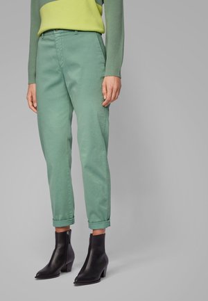 Femme portant un pantalon vert à revers, un haut à manches longues lime et olive, et des bottines noires à talons, debout devant un fond neutre.