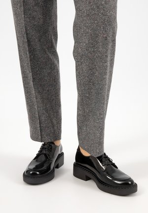 Black patent leather shoes with a thick, textured sole, featuring round toes and laced design, paired with grey patterned trousers.