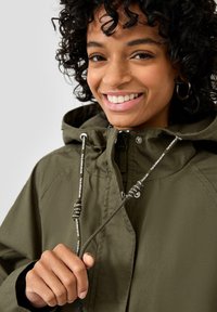 Glunderende vrouw met krullend haar stelt de trekkoorden van de capuchon van haar olijfgroene jas bij.