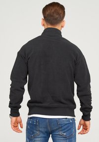 Uomo con capelli corti castani, che indossa una giacca in pile nera, una camicia bianca sotto e jeans blu strappati, in piedi con le spalle alla fotocamera.