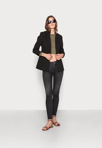 Fekete blézer egy olívazöld crop toppal, sötét skinny farmernadrággal és barna lapos szandállal párosítva. A modell sötét napszemüveget visel, és egy világos háttér előtt áll.