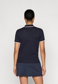 Jūrinės mėlynos spalvos polo marškiniai su kontrastingais baltais apvadais, su tekstūriniais geometriniais raštais nugarinėje pusėje ir prigludusia forma. Trumpomis rankovėmis.