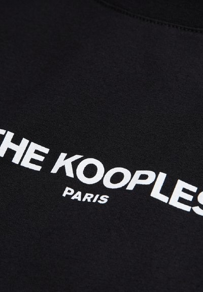 Melna džemperis ar baltu uzrakstu "THE KOOPLES PARIS" modernā, treknā fontā. Uzraksts izvietots diogonāli uz gludas, teksturētas auduma.