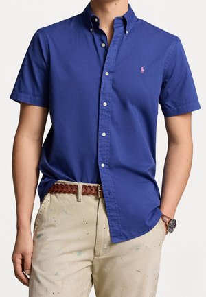 Homme portant une chemise bleue à manches courtes boutonnée avec un logo rose, un pantalon beige éclaboussé de peinture, une ceinture tressée marron et une montre-bracelet.