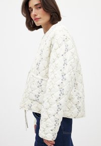 Veste bomber matelassée blanche avec des motifs floraux bleus et verts, offrant une coupe décontractée et un cordon de serrage à l'ourlet.