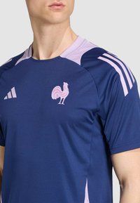 Námornícky modré atletické tričko s fialovými akcentmi a ružovým logom kohúta. Má klasický okrúhly výstrih a tri pruhy na pleciach.