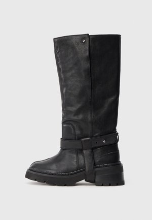 Ψηλή μαύρη δερμάτινη μπότες με χοντρή σόλα, διακοσμητική ζώνη γύρω από τον αστράγαλο και υφή σε αντίθεση με έναν απλό ανοιχτό φόντο.