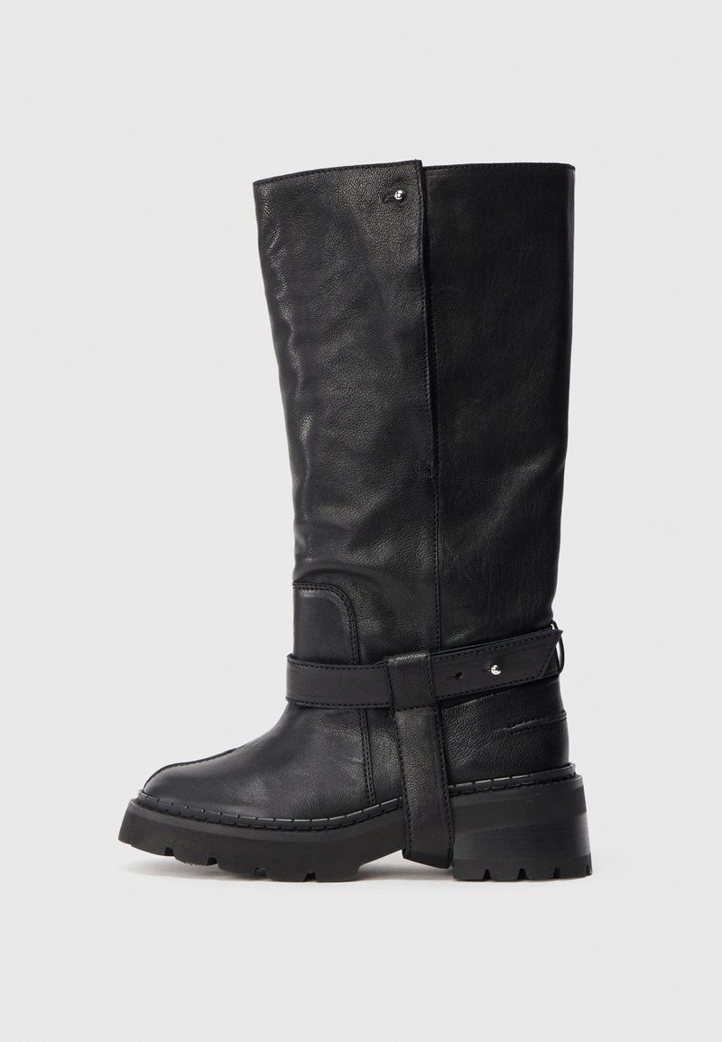 Ψηλή μαύρη δερμάτινη μπότες με χοντρή σόλα, διακοσμητική ζώνη γύρω από τον αστράγαλο και υφή σε αντίθεση με έναν απλό ανοιχτό φόντο.