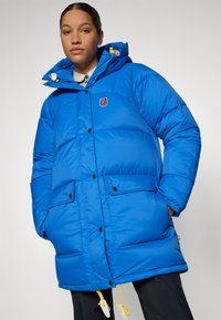 Blauwe pufferjack met een capuchon, voorzien van een logo-embleem, grote zakken en gele accenten. Gemaakt van glad, geïsoleerd materiaal.