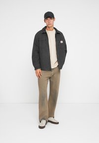 Mørkegrå denimjakke med store frontlommer, beige t-shirt, brune bukser med en falmet effekt, hvide sneakers og en sort kasket.